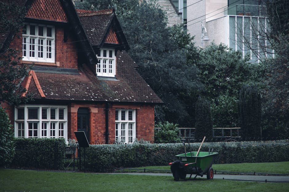 A small front garden featuring a neatly trimmed lawn with vibrant green grass, bordered by a low wooden pathway and a narrow flower bed containing a variety of plants and shrubs. In the background, a 