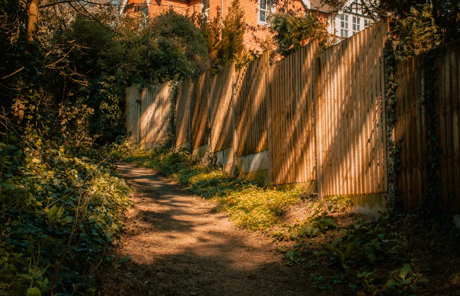 A narrow garden pathway with a compacted and slightly uneven dirt surface, bordered by dense green shrubbery and low-lying plants on the left. On the right, there is a tall wooden fence made of vertic